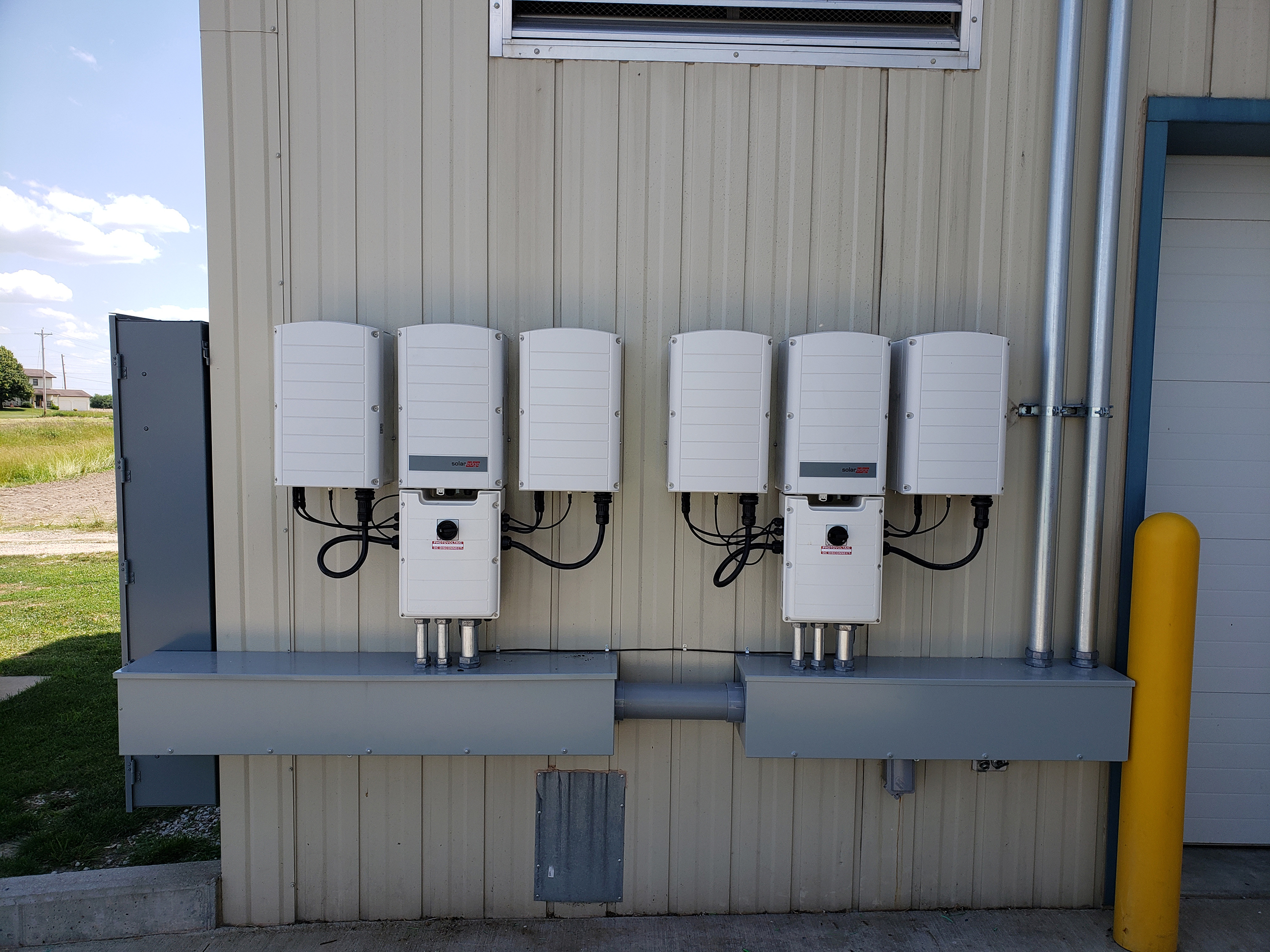 The corner of a tan metal building has six boxes neatly lined up with organized wiring and conduit running between the systems. The entrance of a garage door is seen to the right.