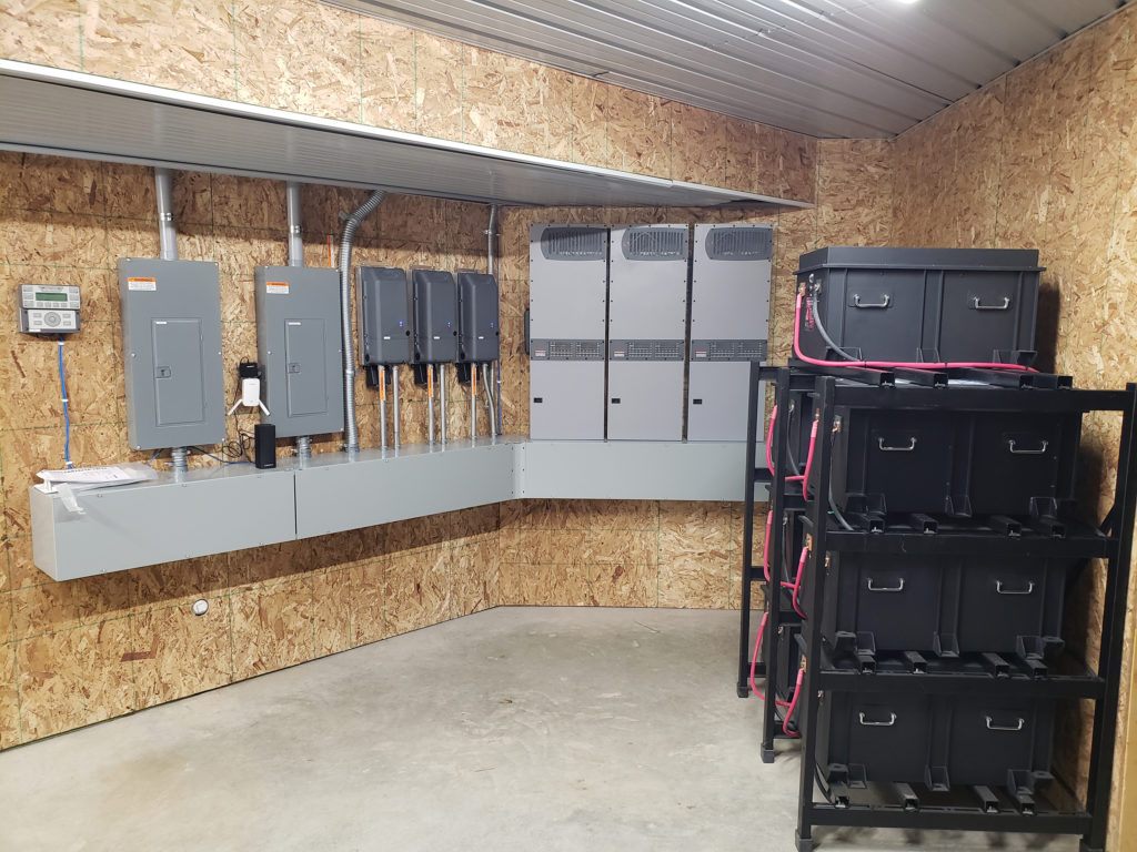 The components of the battery system, energized by solar energy, is neatly organized in an unfinished room. The electric panels and wiring boxes neatly line the left wall. A rack with large batteries is seen on the right.