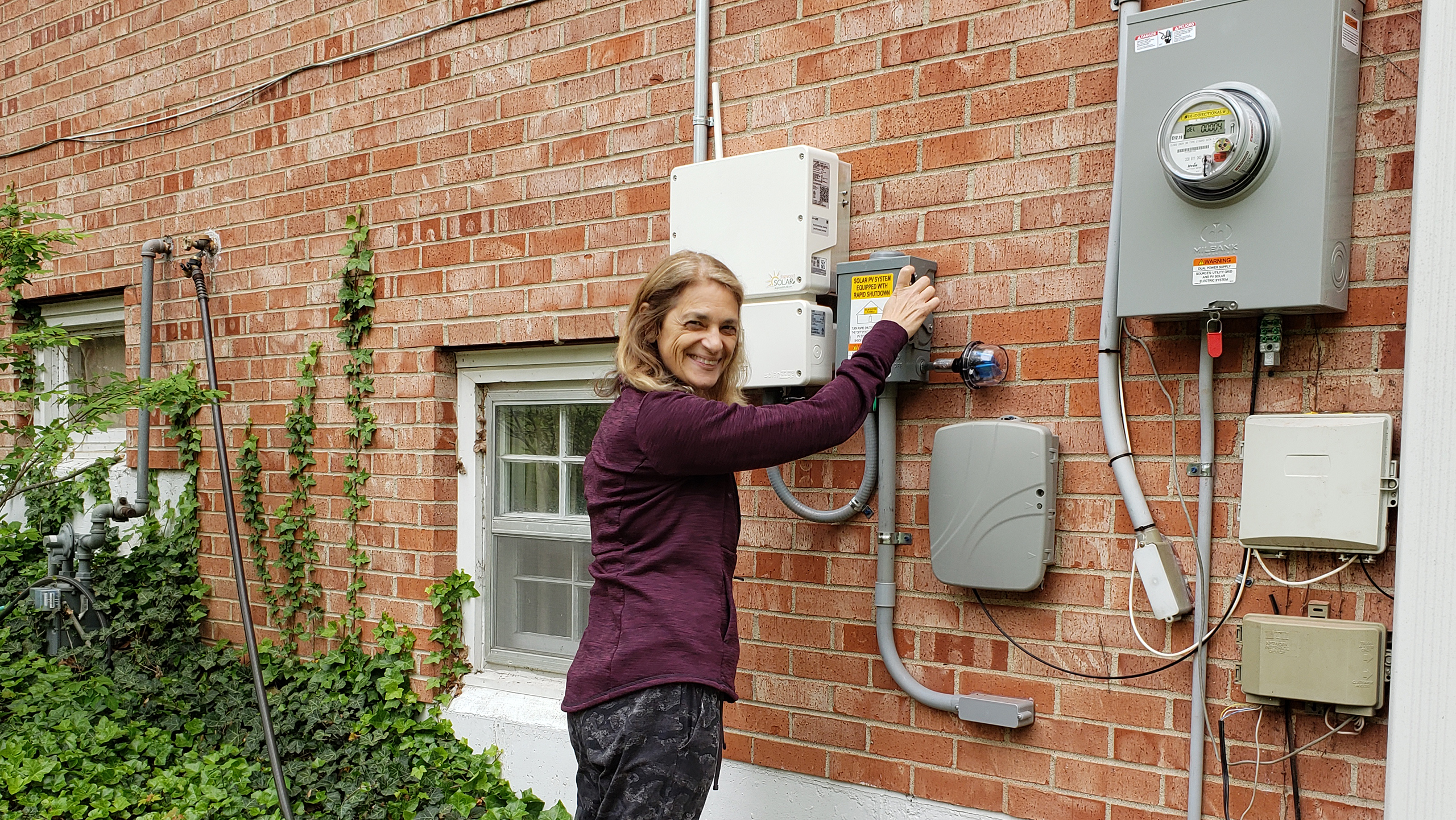 Woman stands posed to flip the switch where the solar system integrates with the utility panel outside a brick home. She is smiling and looking over her shoulder at the camera. Some green vines can be seen to her left, showing it is an established home.